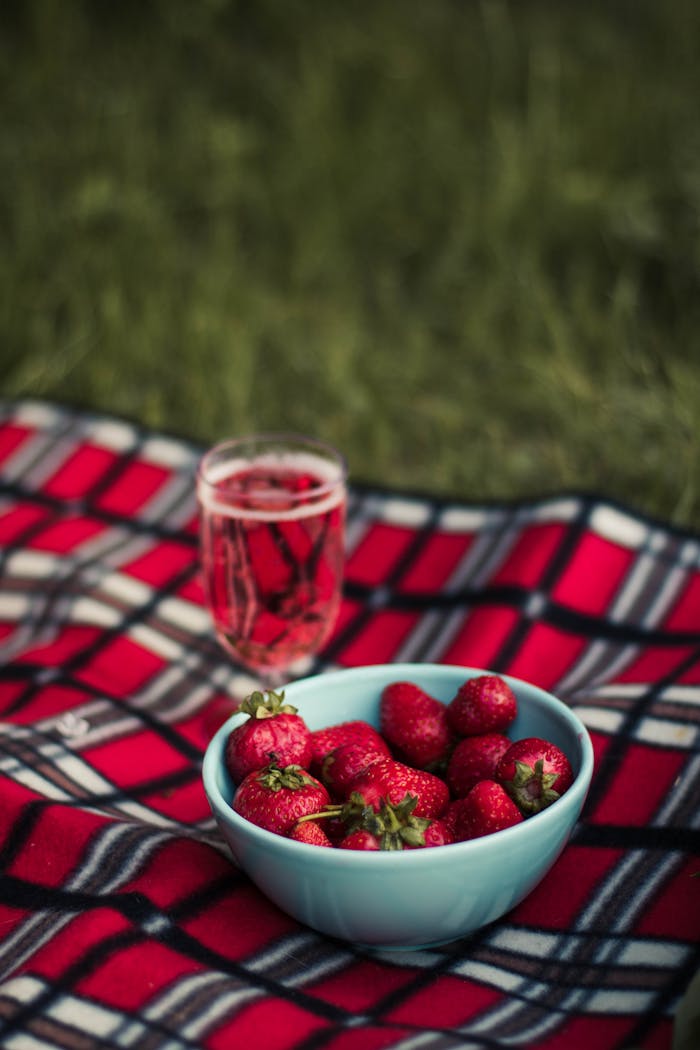 A bowl of ripe strawberries on a plaid picnic blanket with a drink, set outdoors.